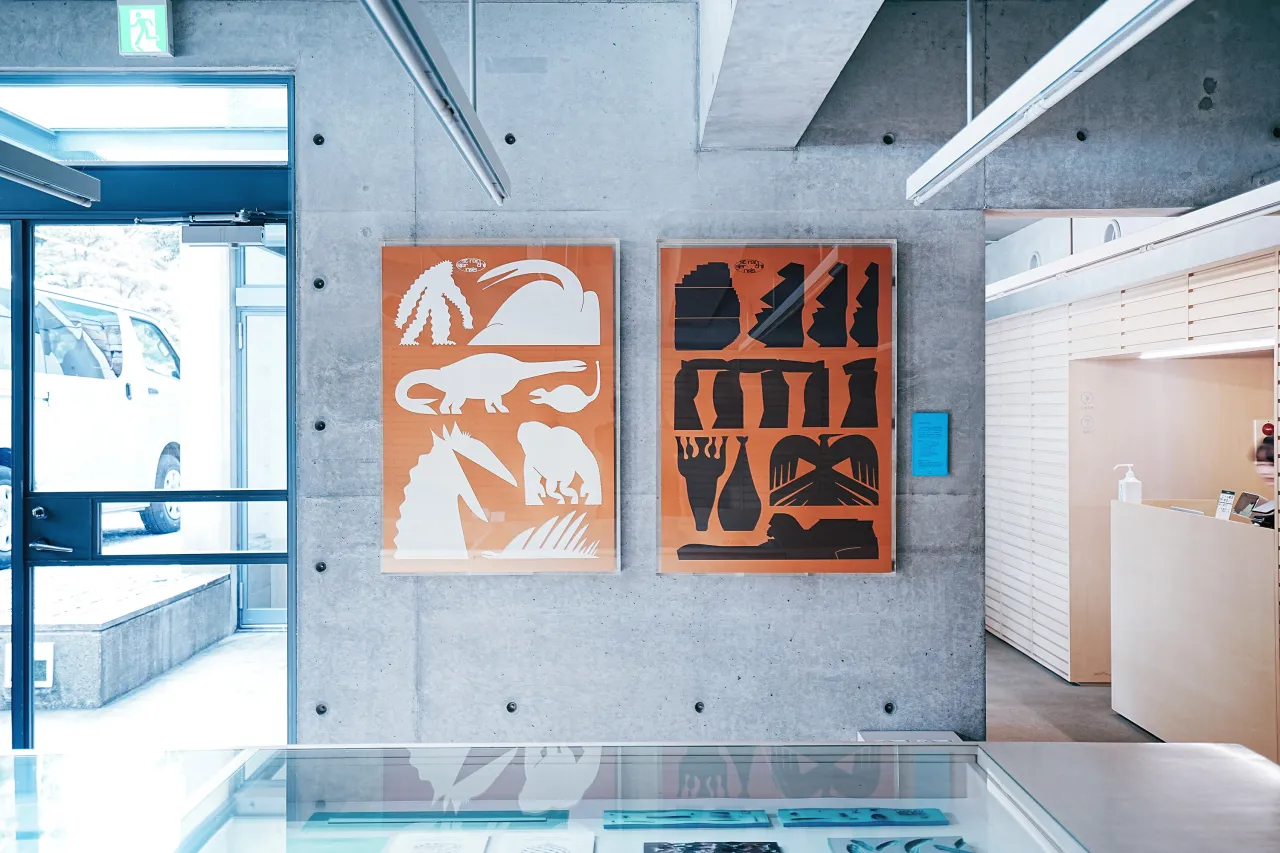 Two orange paper-cut artworks depicting stylised animals and abstract shapes hang on a concrete wall in a modern office space with blue-framed glass doors and white reception desk